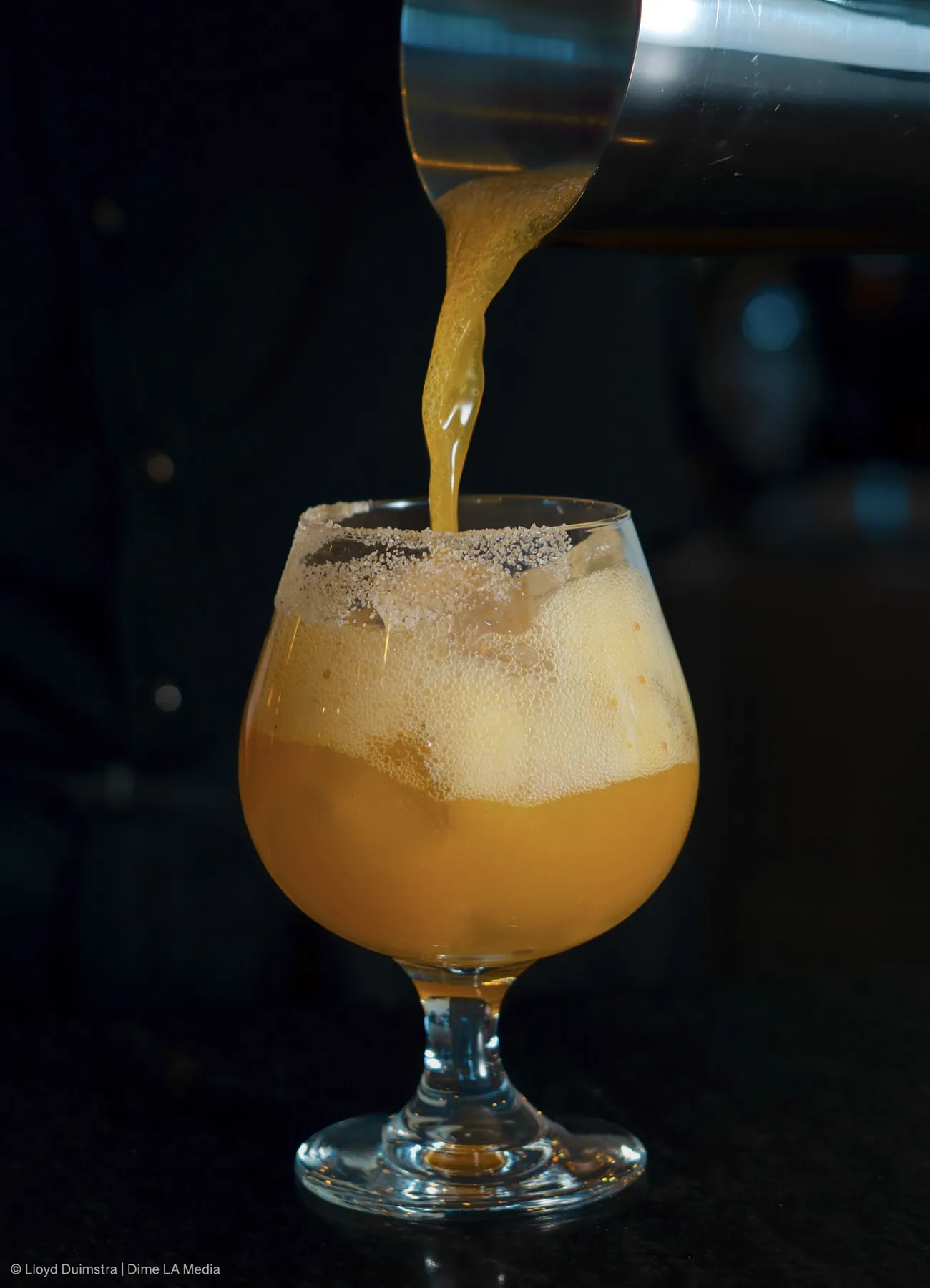 Amber cocktail with a sugared rim being poured from a shaker into a snifter glass in low light