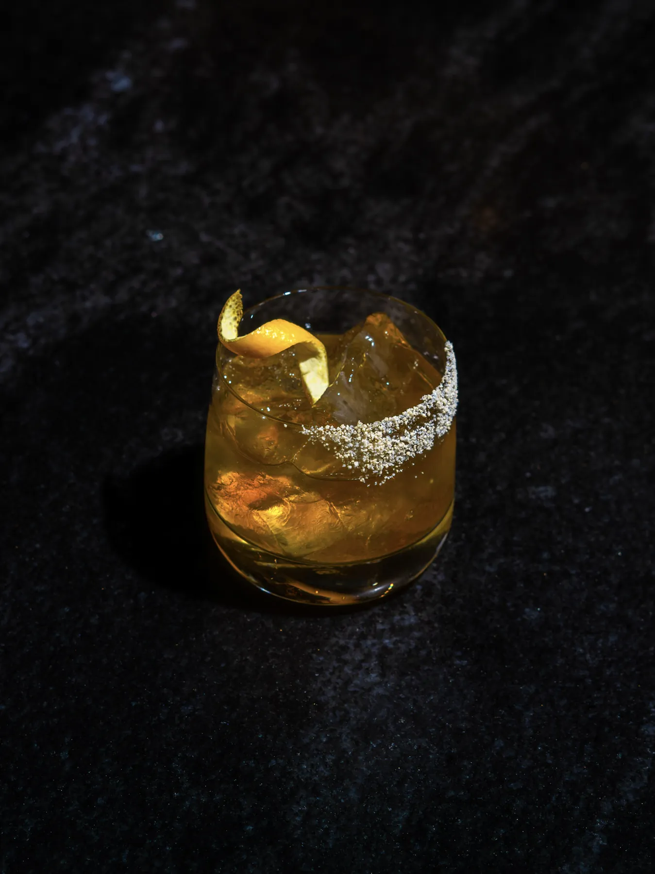 Whiskey cocktail on the rocks with a citrus twist and sugared rim on dark stone