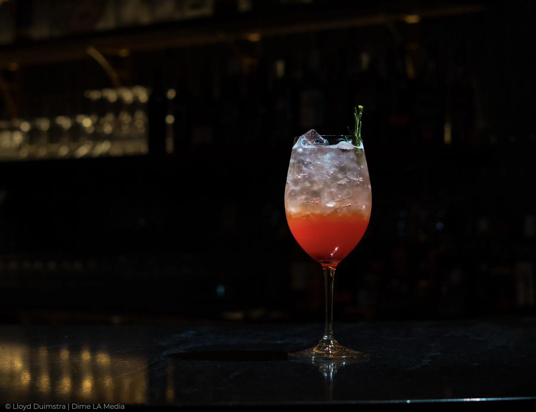Red ombre spritz with a rosemary garnish in a dim, atmospheric bar