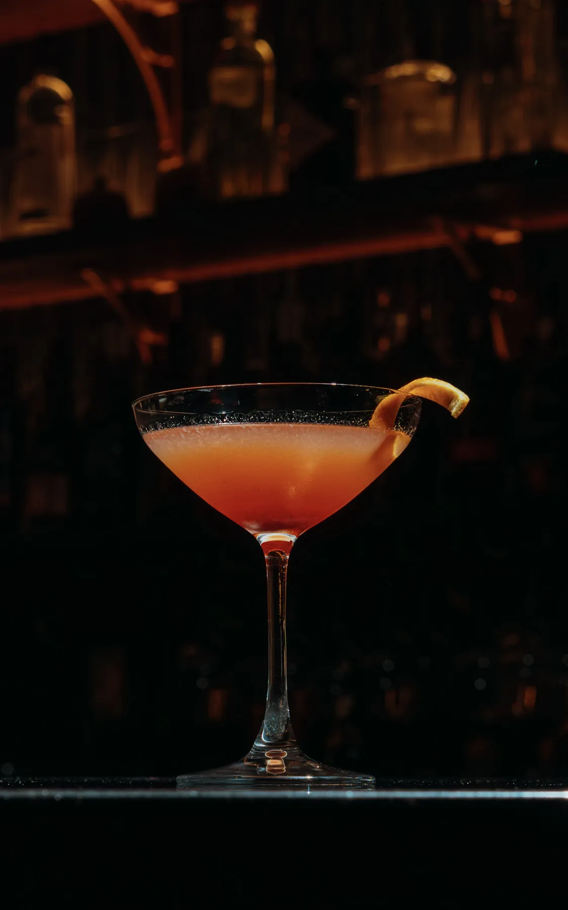 Manhattan-style cocktail in a coupe with an orange twist in low light