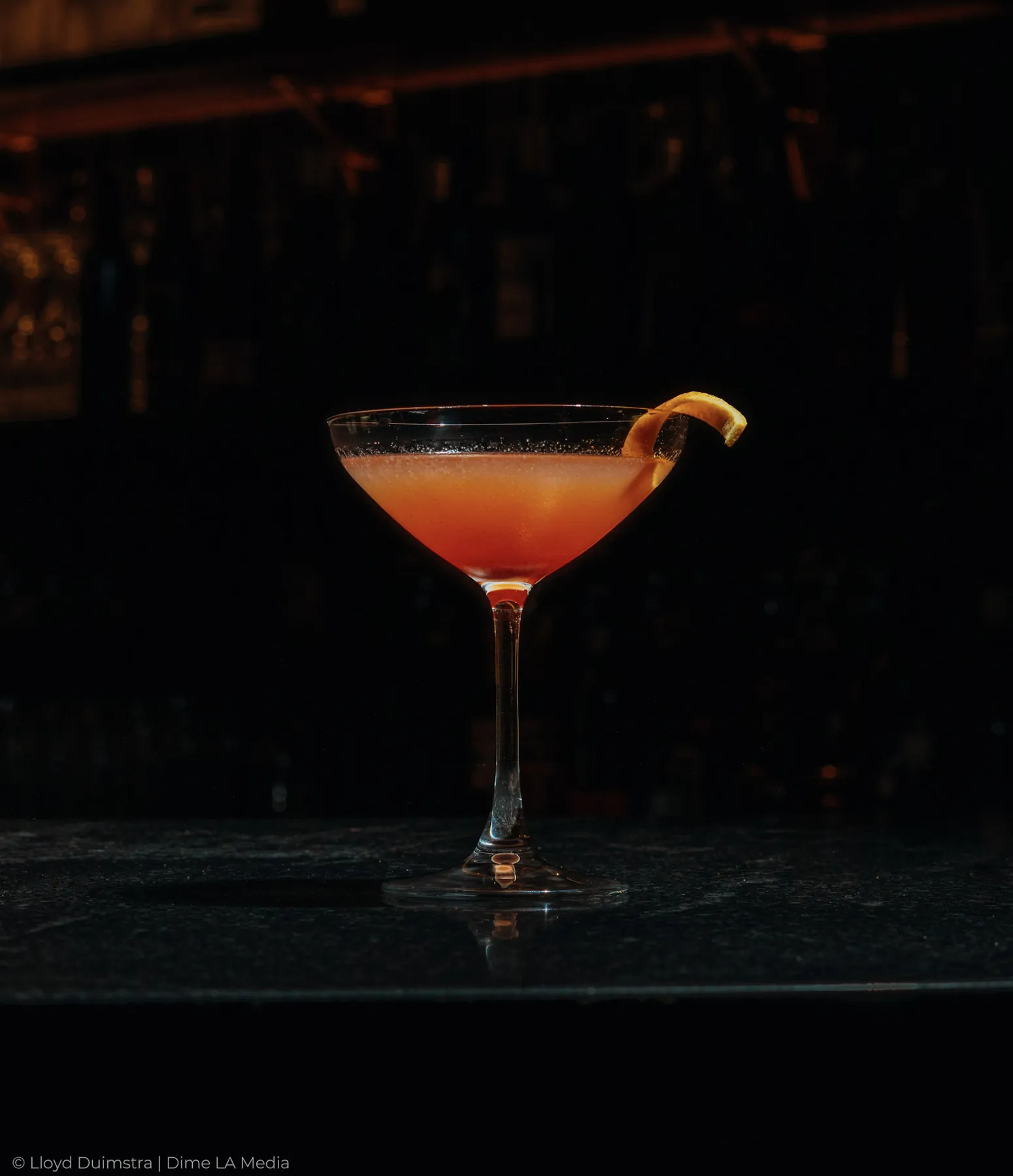 Amber coupe cocktail with a citrus twist against the back bar