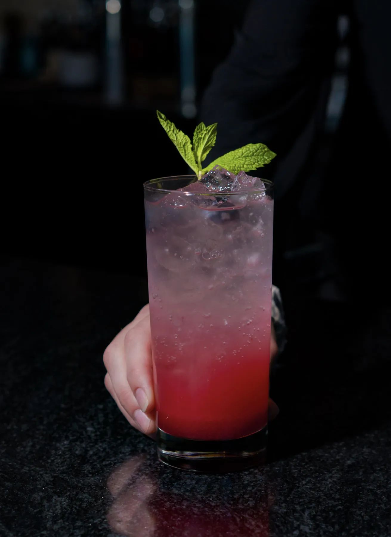 Tall pink ombre cocktail garnished with mint, held on a dark bar top