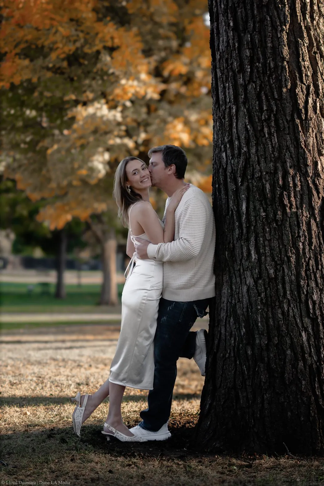 Man kissing his fiancee's cheek beneath an autumn tree