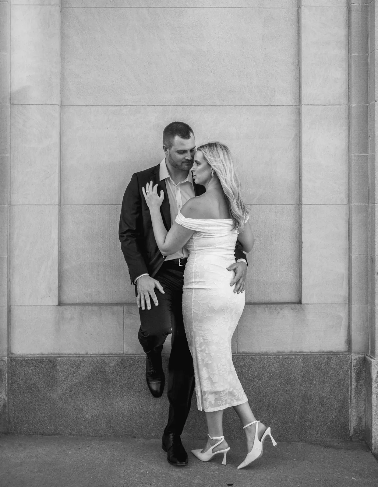 Black and white full-length portrait of an engaged couple against a carved stone wall