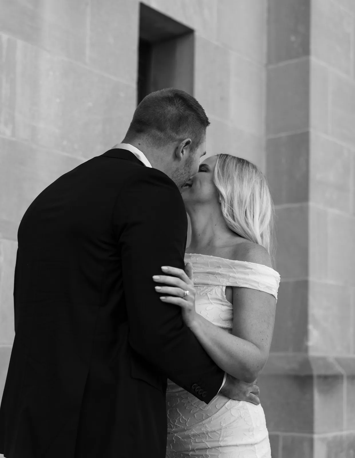 Black and white photo of an engaged couple kissing against the cathedral limestone