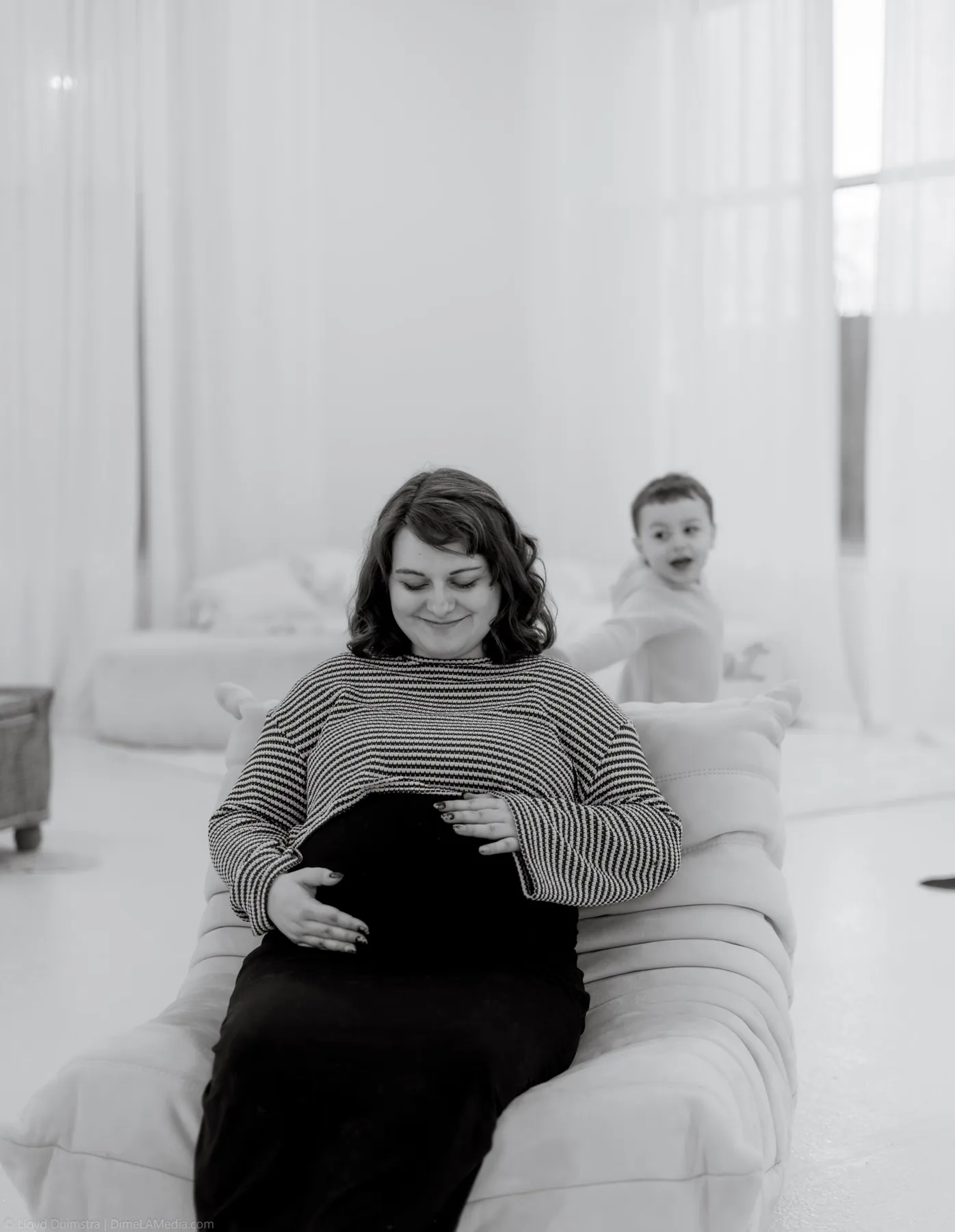 Black and white maternity portrait with the toddler peeking behind the couch