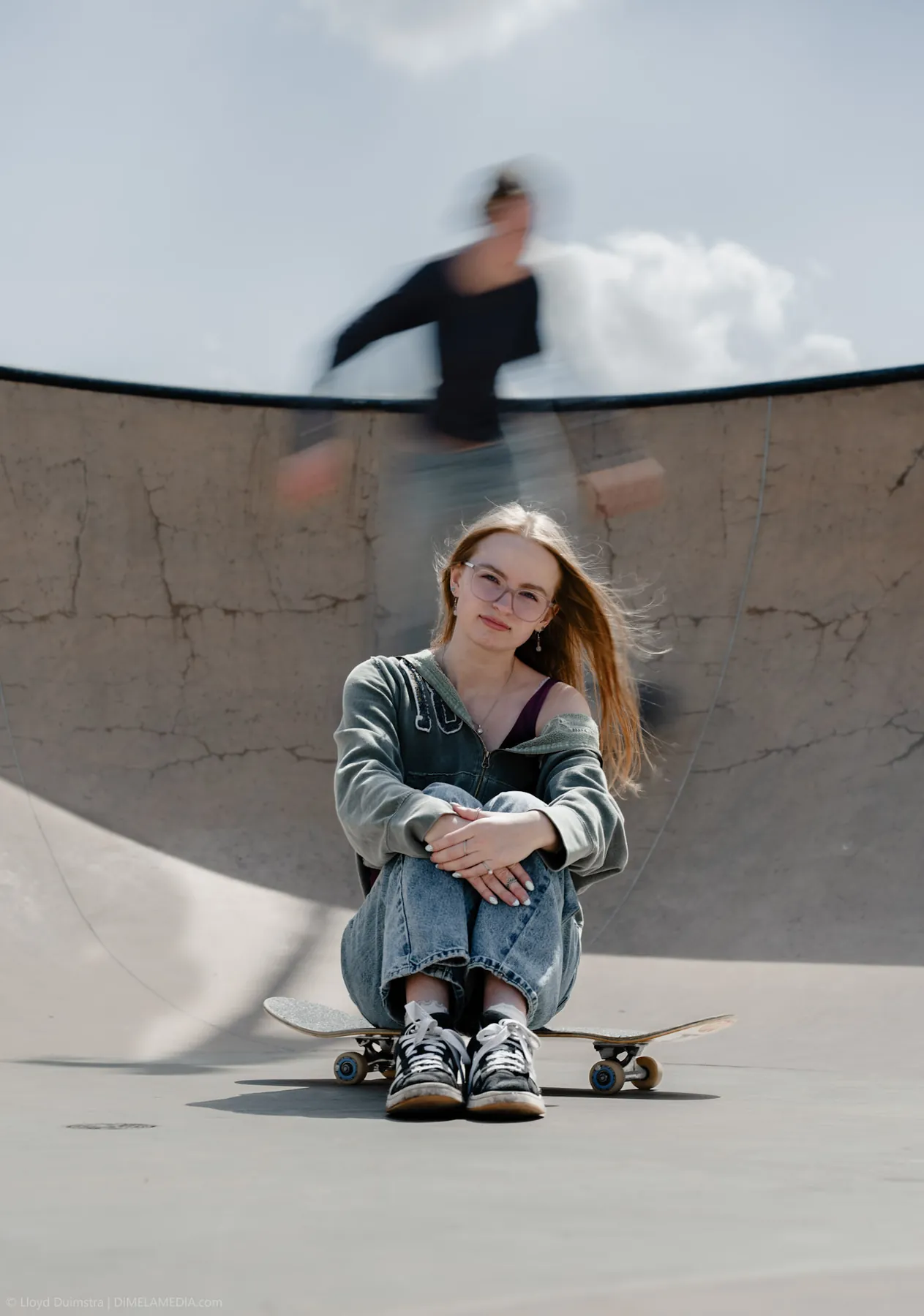 Syd Patzwald sitting on her skateboard at the skatepark as a skater blurs past
