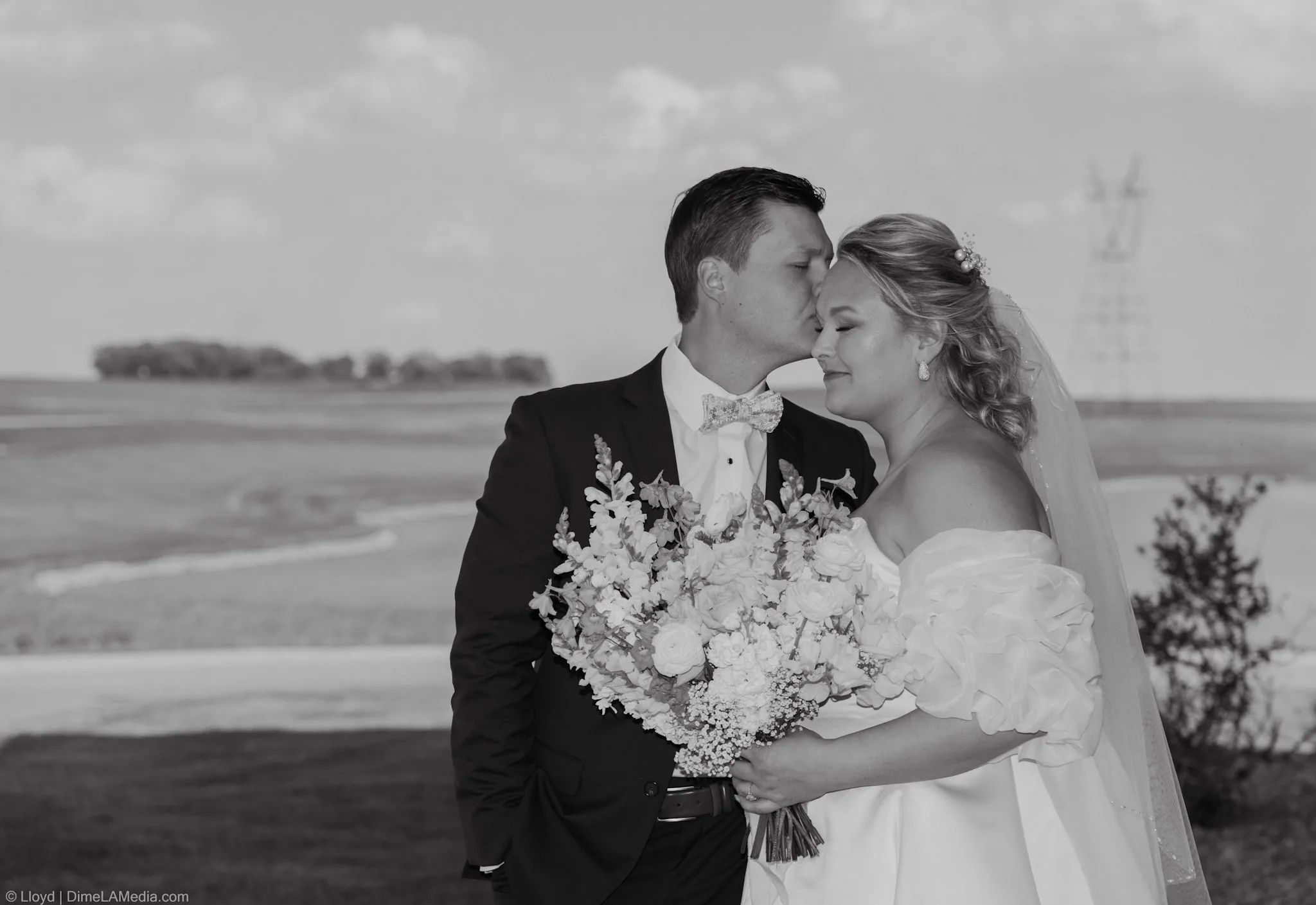 Groom kissing his bride's forehead over her bouquet, black and white