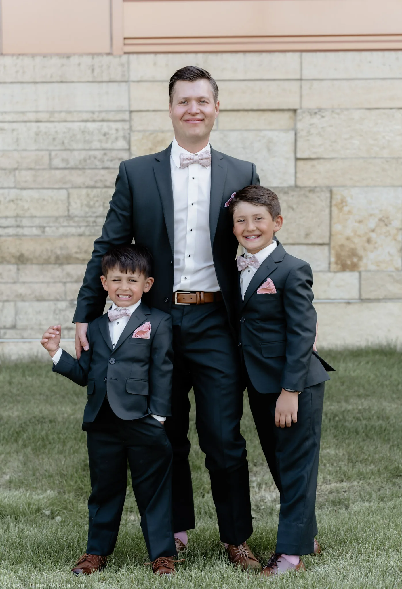 Groomsman with two young attendants in matching charcoal suits and blush bow ties against a limestone wall