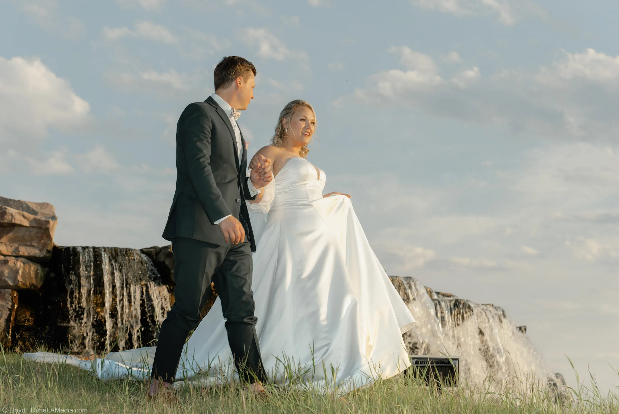 Bride lifting her dress as she and her groom walk through the grass beside the falls